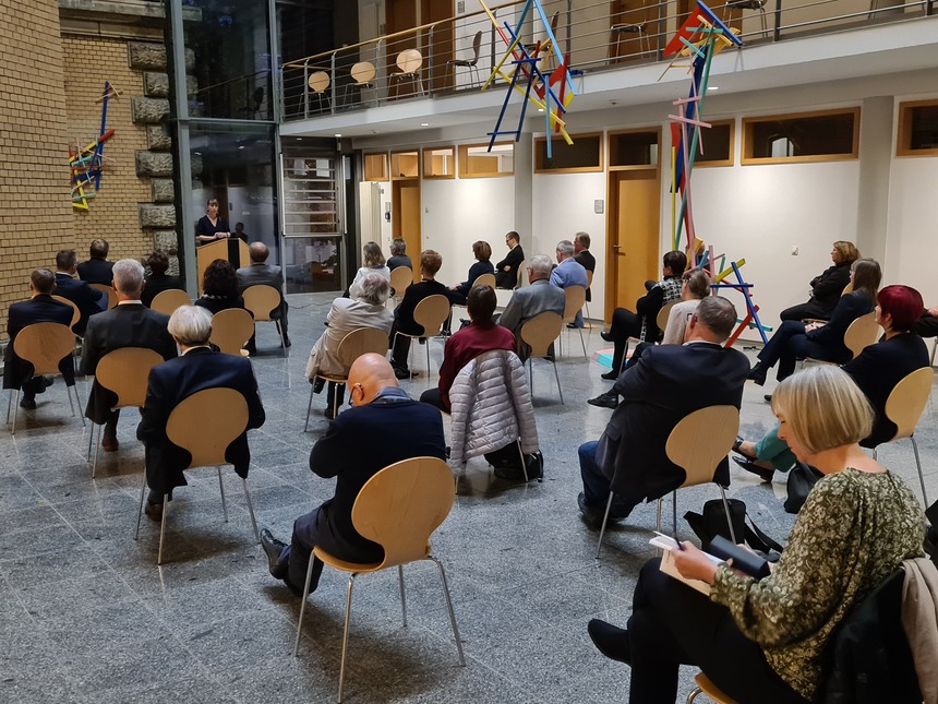 Menschen sitzen in großen Abständen voneinander im Lichthof des Justizministeriums, die Ministerin steht ganz hinten vor einem Rednerpult und spricht.