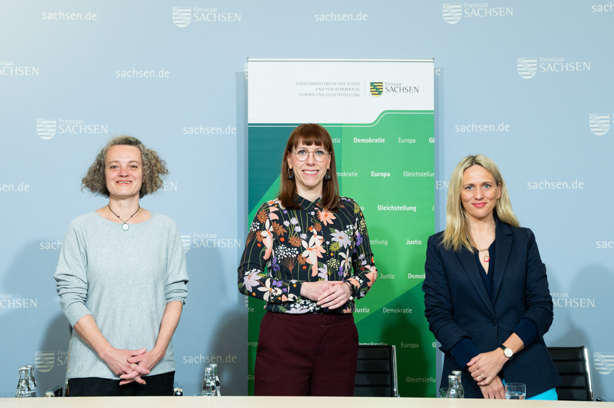 Susanne Burkhardt, Justizministerin Katja Meier und Prof. Dr. Elisa Hoven stehen vor einer Pressewand des Freistaates Sachsen und des Sächsischen Staatsministerium der Justiz und für Demokratie Europa und Gleichstellung. Beides steht auch im Hintergrund.