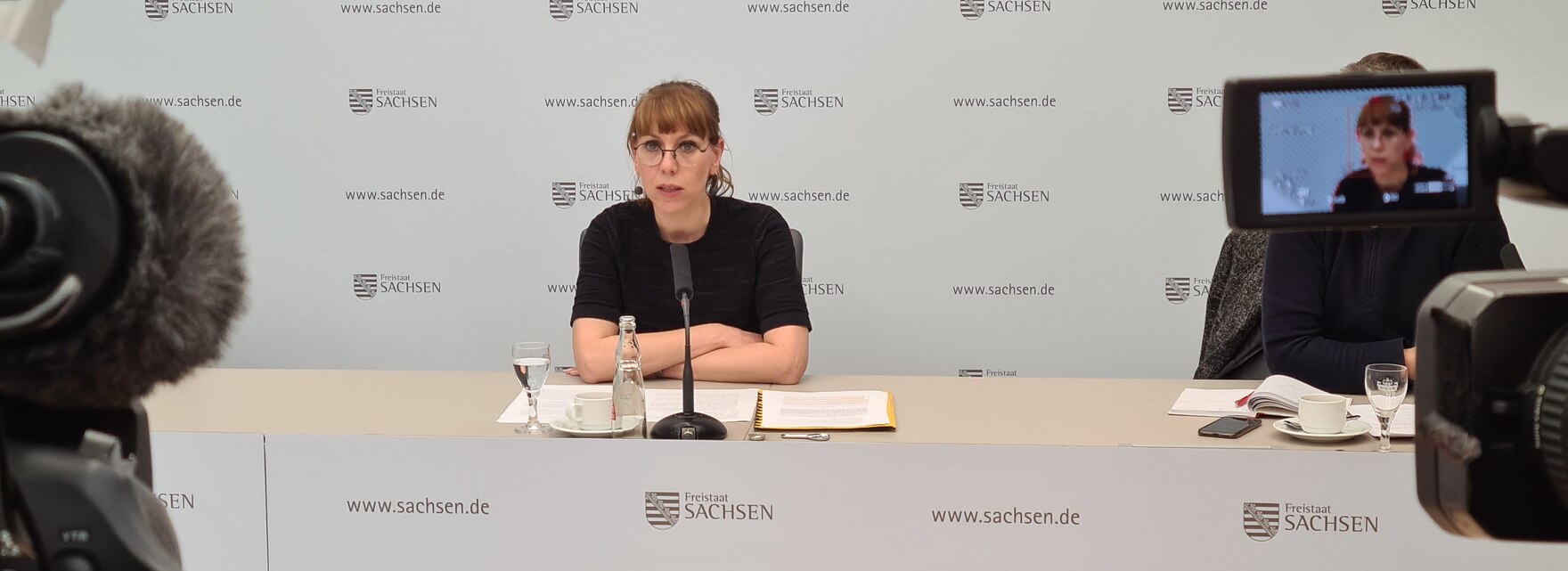 Katja Meier mit verschränkten Armen an einem Tisch, im Hintergrund eine weiße Wand mit Schriftzügen wie Freistaat Sachsen etc.