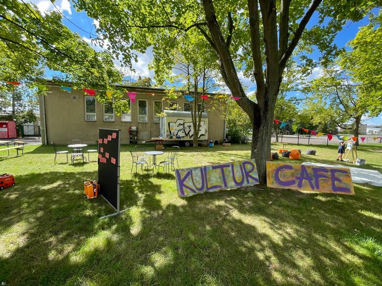 Wiese mit Baum und Gebäude im Hintergrund, im Vordergrund ein selbstgesprühtes Bild mit der Aufschrift KULTURCAFE