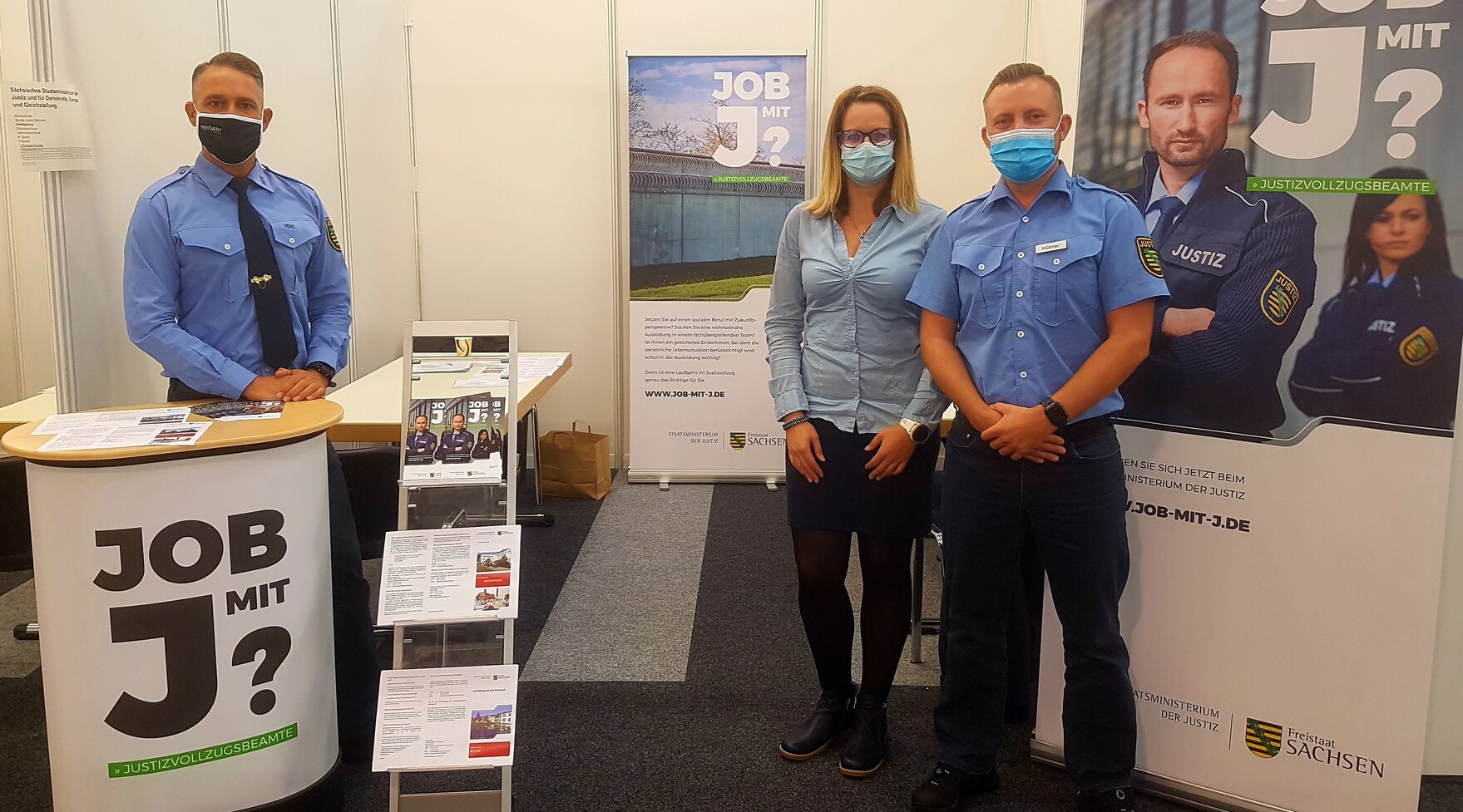 Eine Frau und zwei Männer in blauen Uniformen an einem Messestand, An der Seite links Plakat "Job mit J"