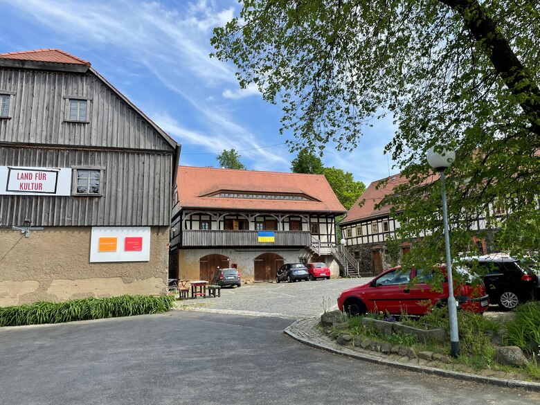 Begegnungszentrum in einem Dreiseithof. Im Vordergrund an der Stirnseite eines Hauses ein Banner: "Land für Kultur". An der Brüstung eines Hauses im Hintergrund hängt die Ukraine-Flagge.